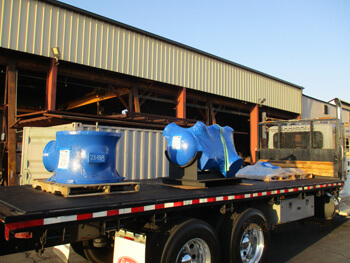 Flatbed truck carrying large blue industrial components outside a metal fabrication facility in daylight.