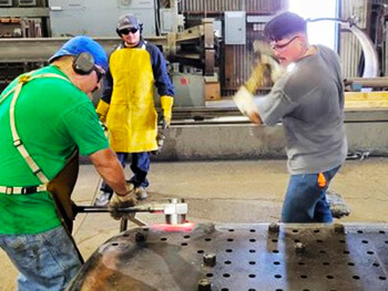 Industrial workers using hammers and tools to assemble or repair a large metal heat exchanger tube sheet in a fabrication workshop.