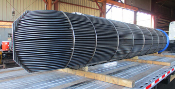 Large cylindrical heat exchanger tube bundle with metal support rings, placed on a wooden platform inside an industrial workshop.